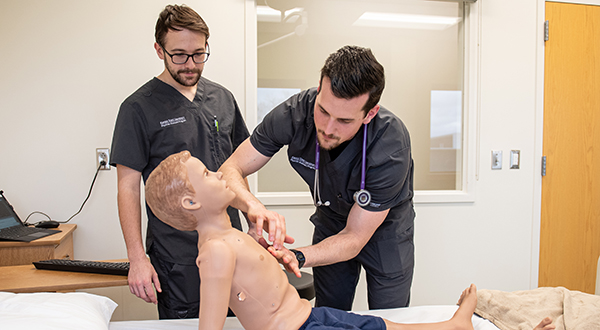 Two male students in dark gray scrubs practice their examination skills on an advanced patient simulator that looks like a male child. One student who is wearing a purple stethoscope places his hands on the patient simulator's chest.