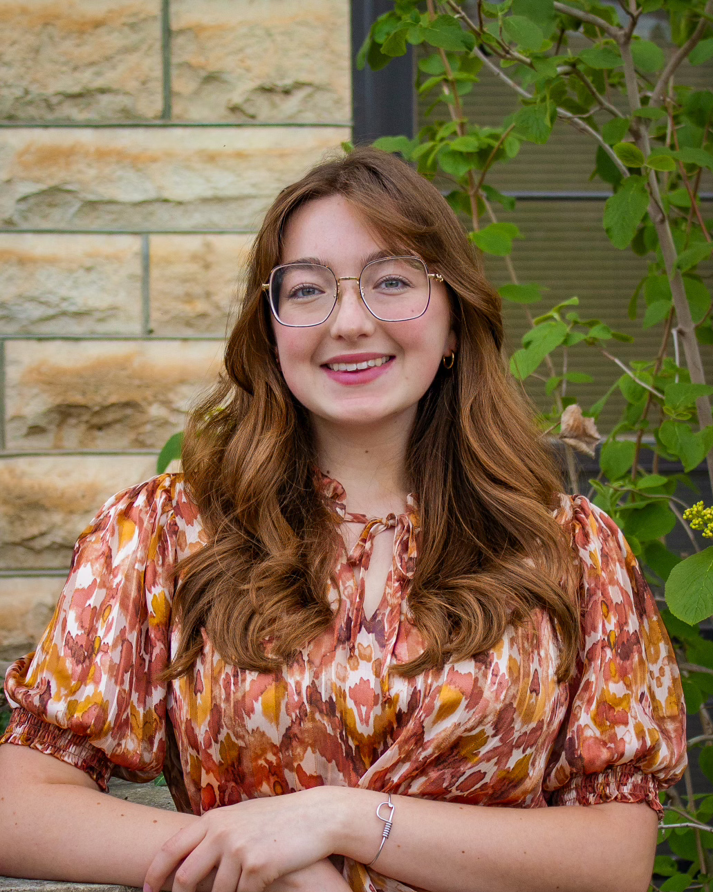 A young woman with glasses and a red and orange patterned shirt smiles for a photo.