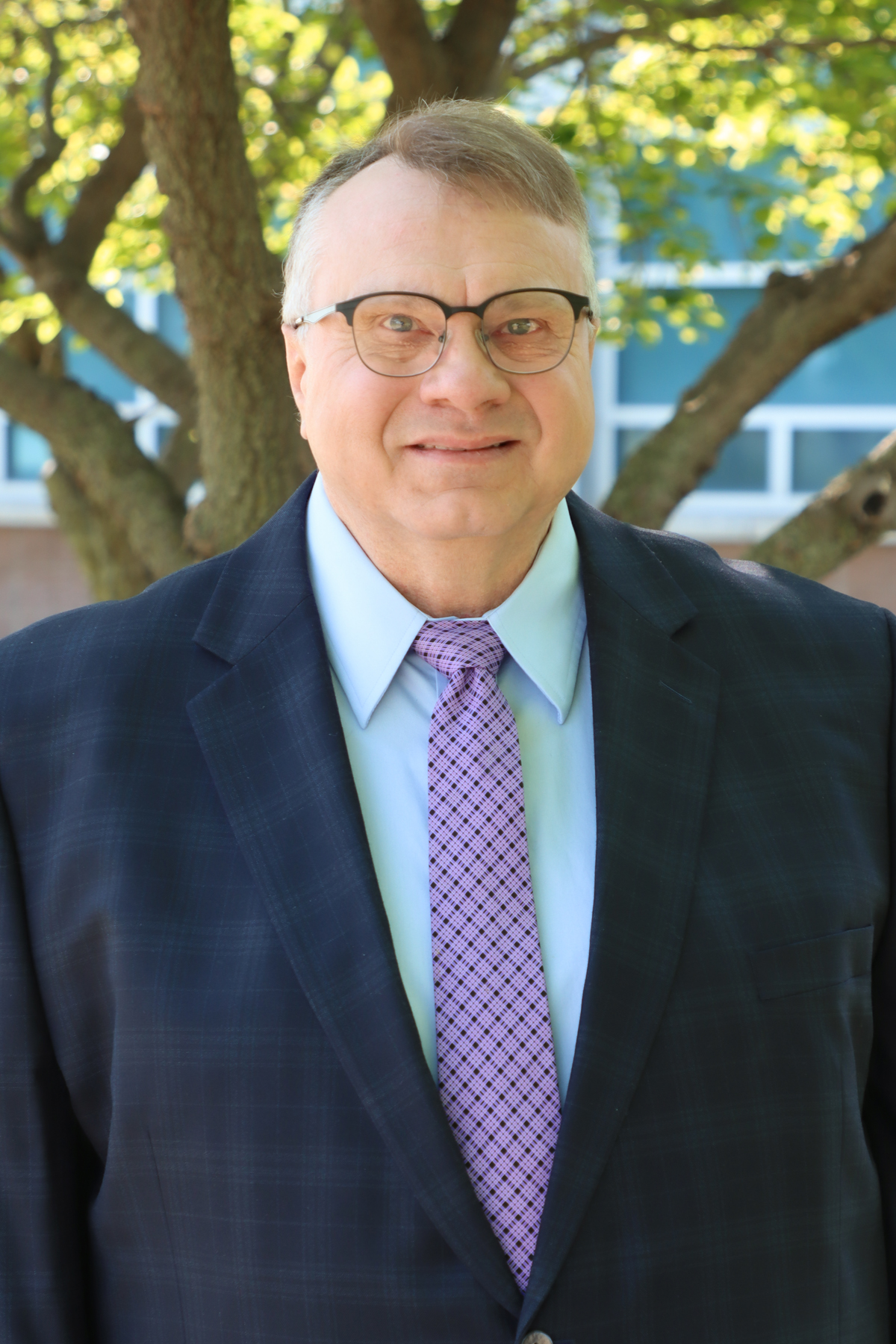 A white man in glasses and a blue suit with a purple tie smiles for a portrait photo.