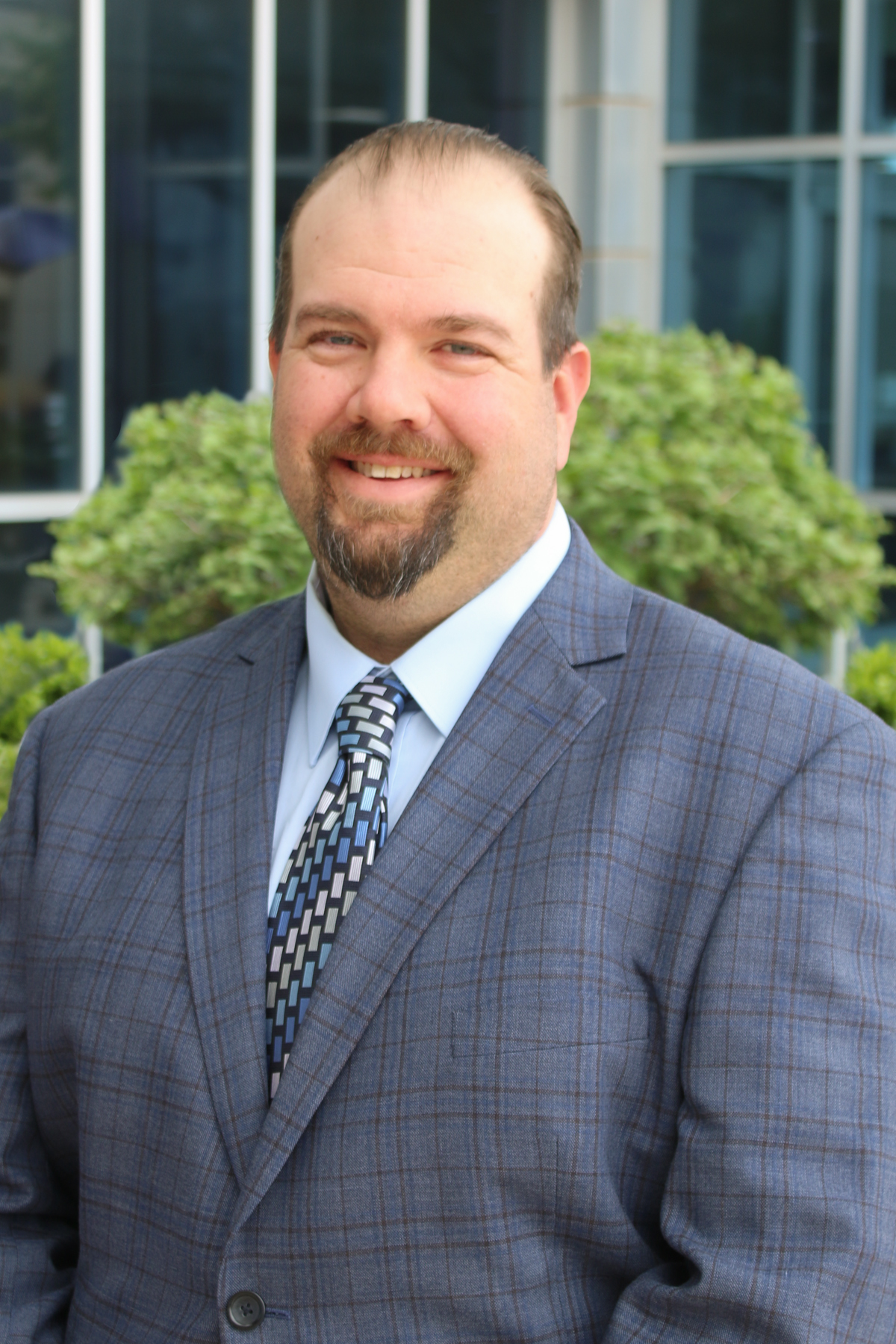 A white man with brown hair in a blue patterned suit and tie smiles for a portrait photo.