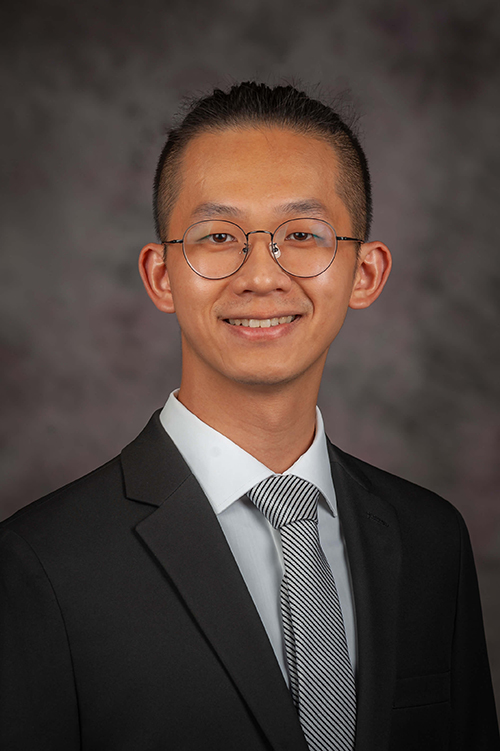 A student with round glasses in a suit and tie smiles for a portrait photo.