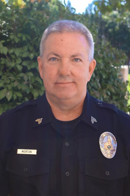 A white man in a police uniform with a badge and nametag that reads Morton smiles for a portrait photo.