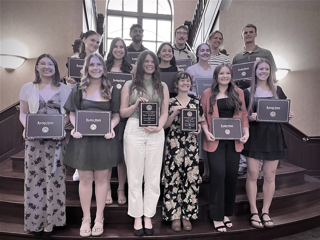A group of undergraduate biology students poses with award placards on a wooden staircase.