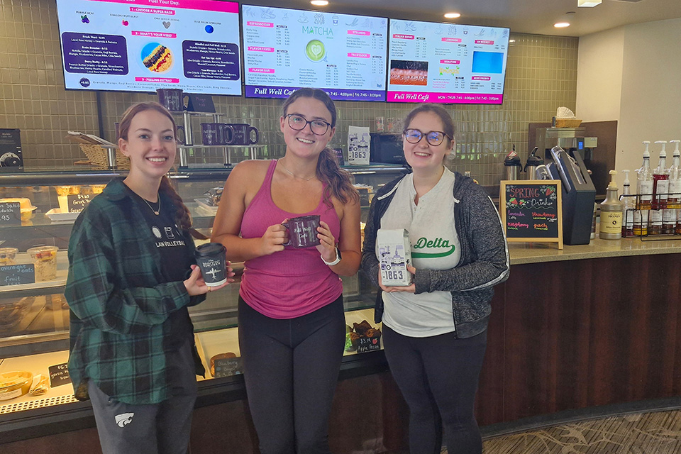 Three students pose for a photo in a café holding café items. 