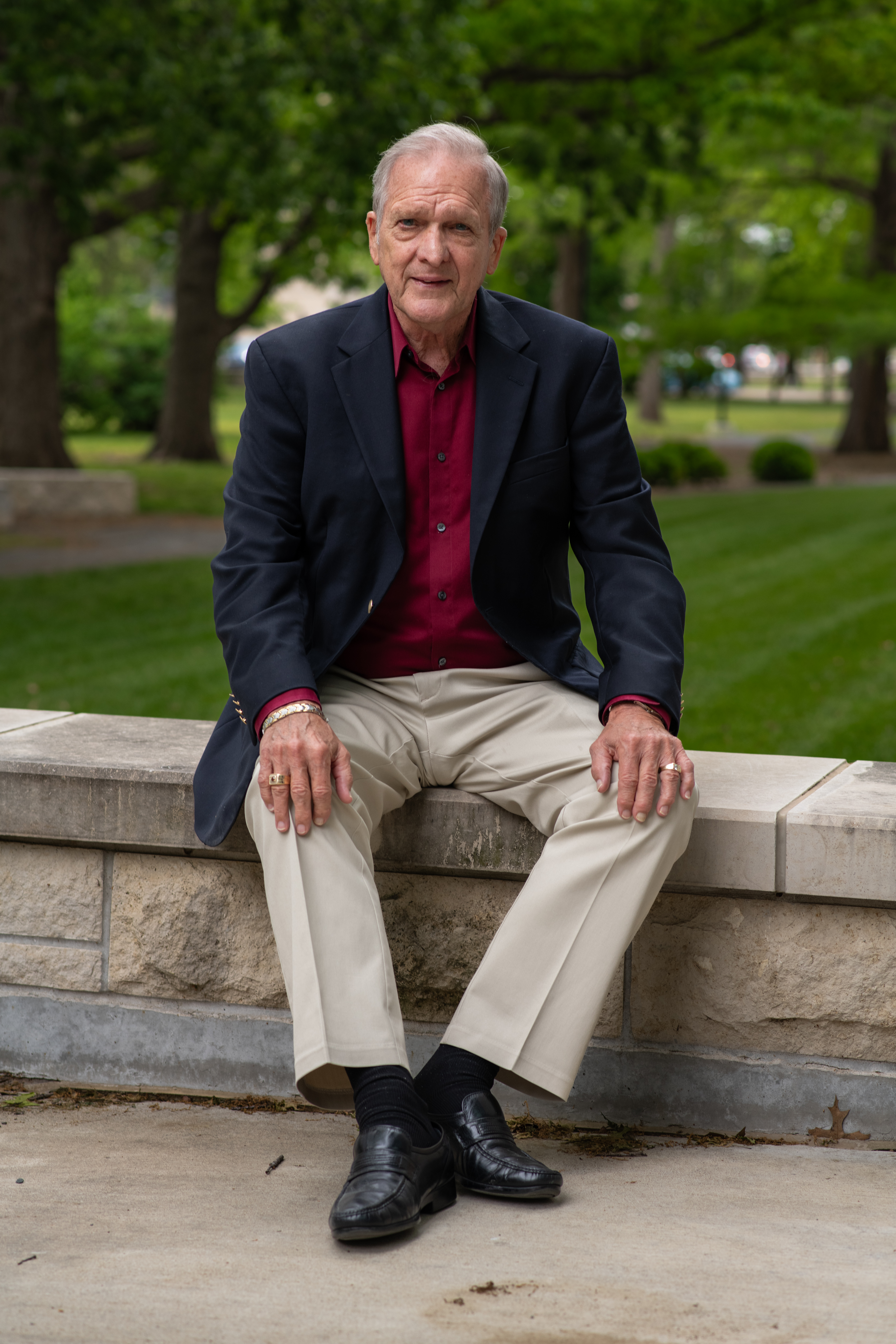 A gentleman in a red shirt, black blazer and khaki pants sits on a low limestone wall against a green background of trees and grass.