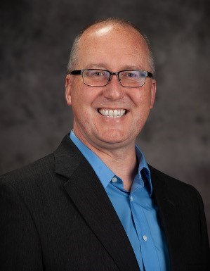 Man in a blue button-up shirt, black blazer and glasses smiles against a gray backdrop for a portrait. 