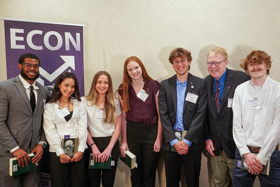 Seven people in business attire stand in a row for a group photo in front of a purple banner with the word Econ written on it. 