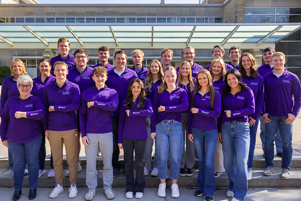 A group of students in purple polo pose for a group photo outdoors in front of the K-State Student Union.