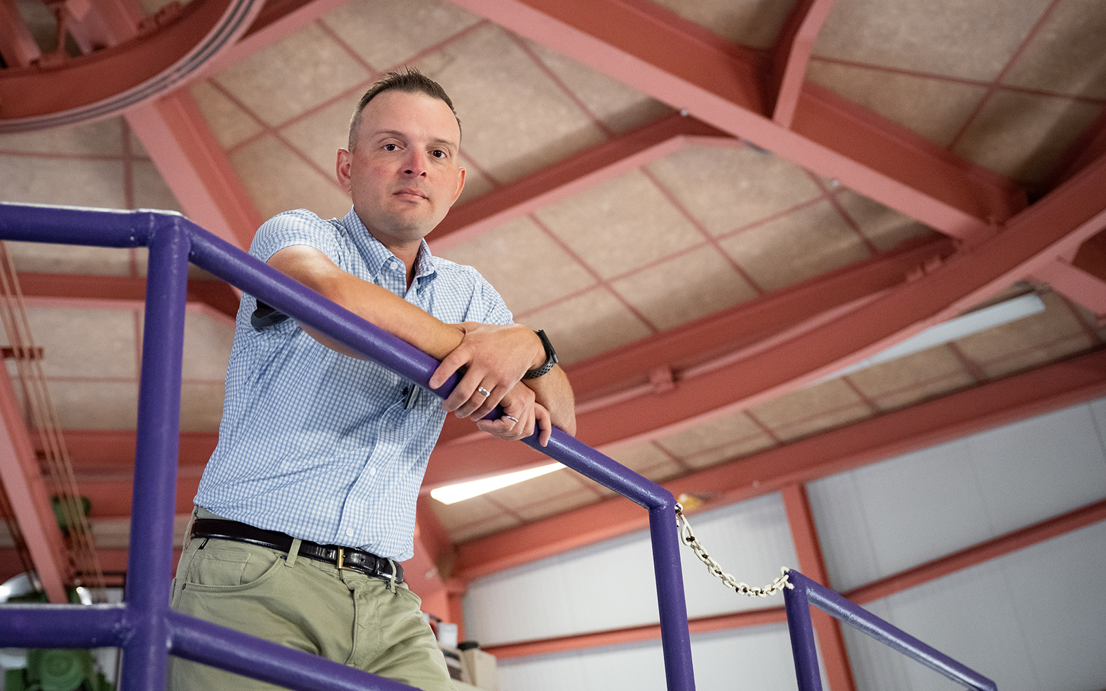 A college nuclear engineering professor poses for a portrait in a large reactor room while leaning forward on a purple rail.