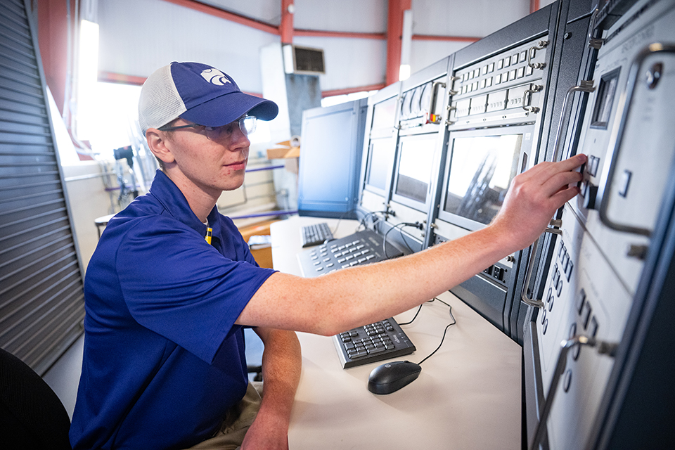 A student in purple polo and ball cap works with panels at a nuclear reactor console.