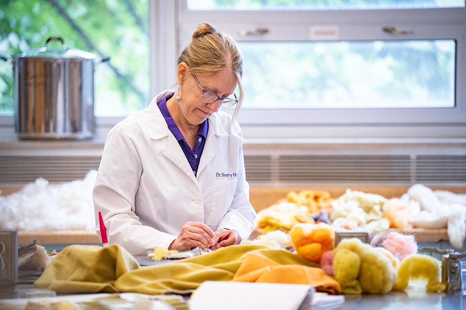 A woman in a white lab coat works with various dyed fabrics in a lab with windows.