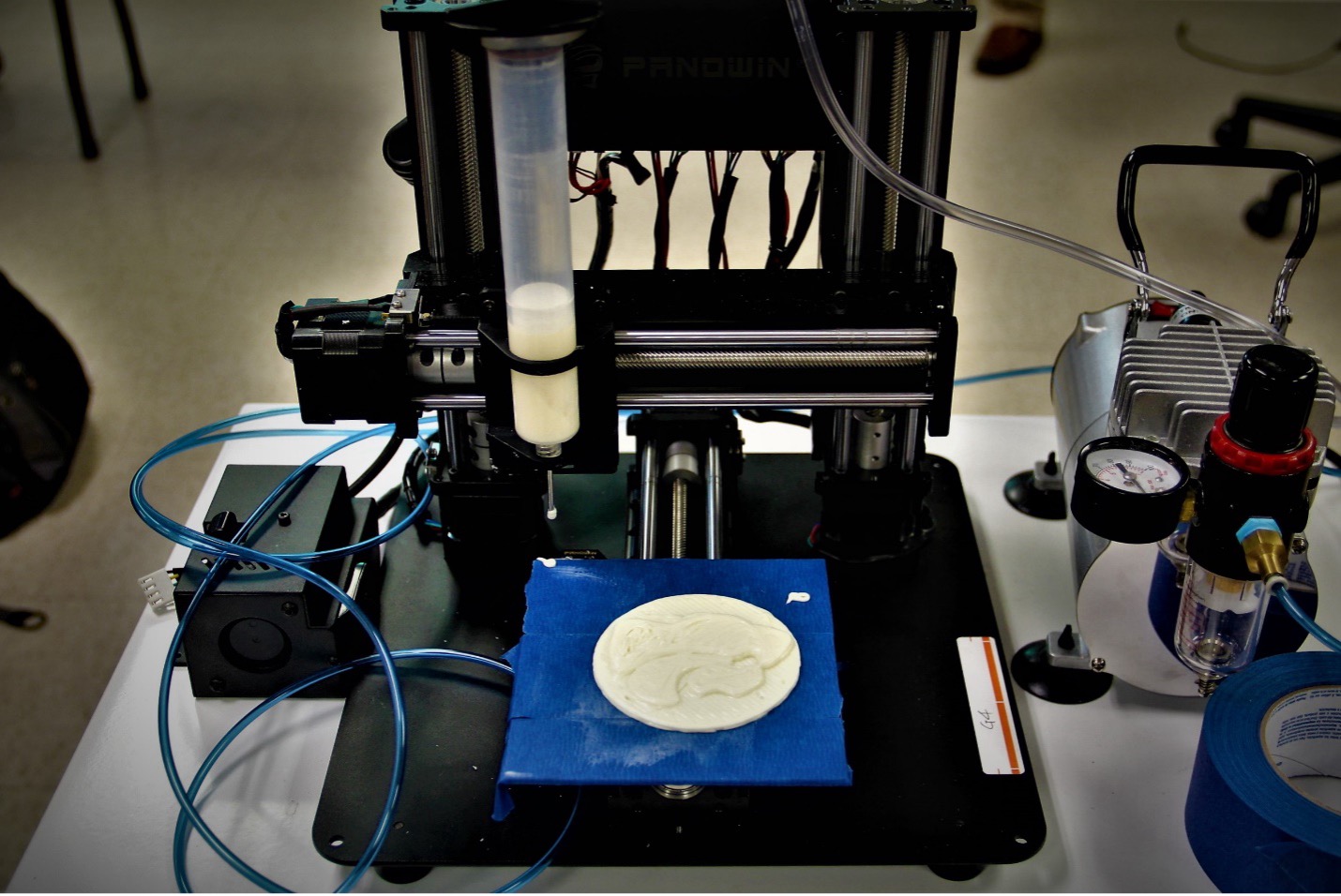 A white syringe filled with a white goo hangs on machinery above a white circle of the goo that itself is on several strips of blue cloth. The white circle of goo has in its center a built-up formation of the K-State Powercat logo.
