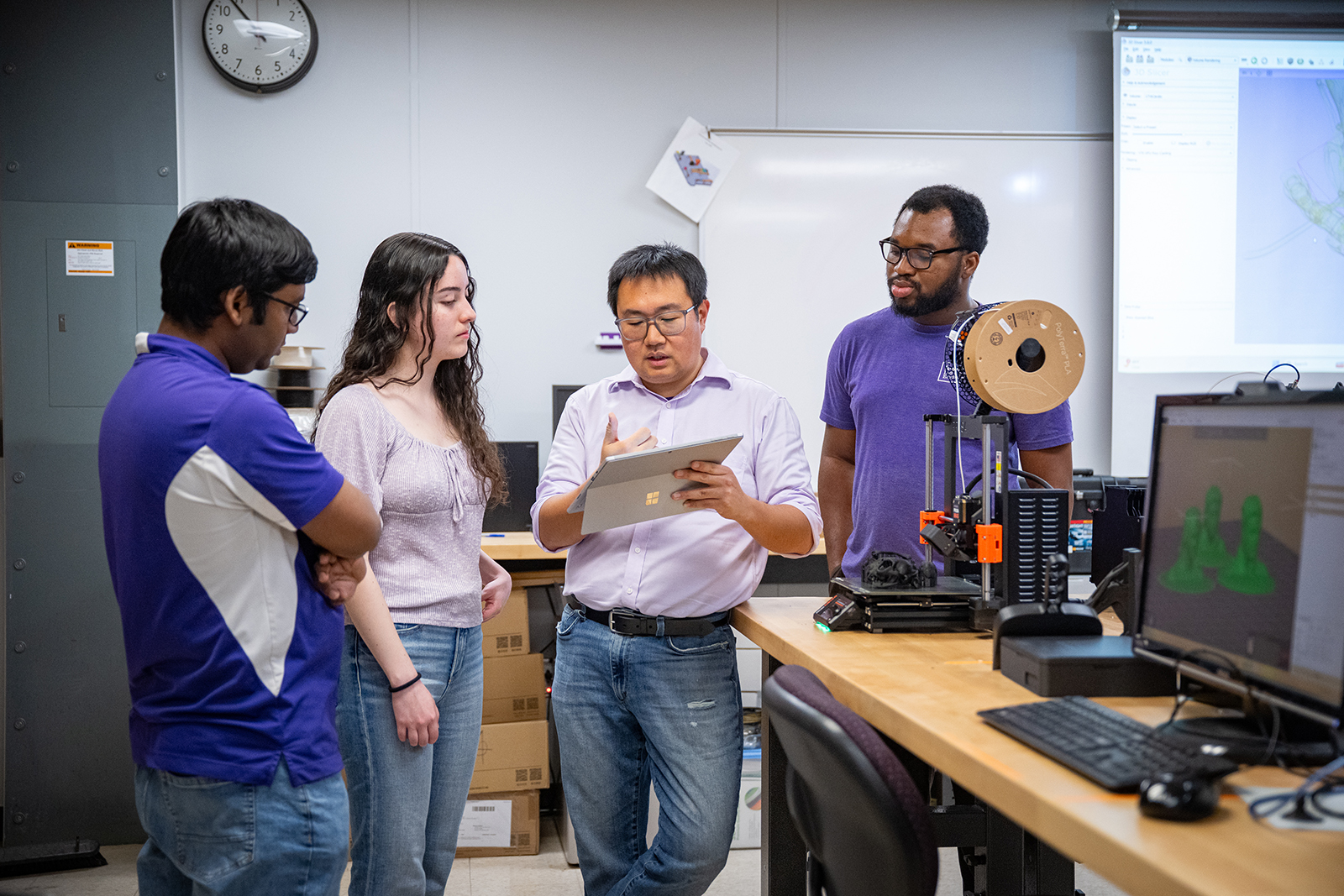 In an engineering lab, a professor holds up a tablet computer and explains concepts to a group of three undergraduate students who listen around him.