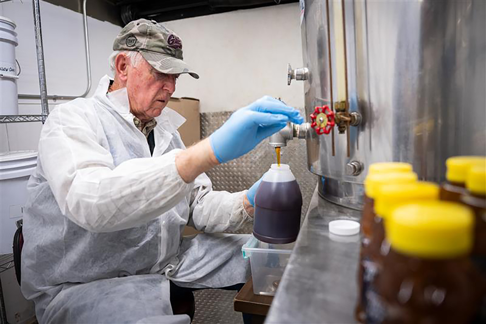 A man in a white coat pours honey into a jug out of a cylindrical metal container with a spigot.