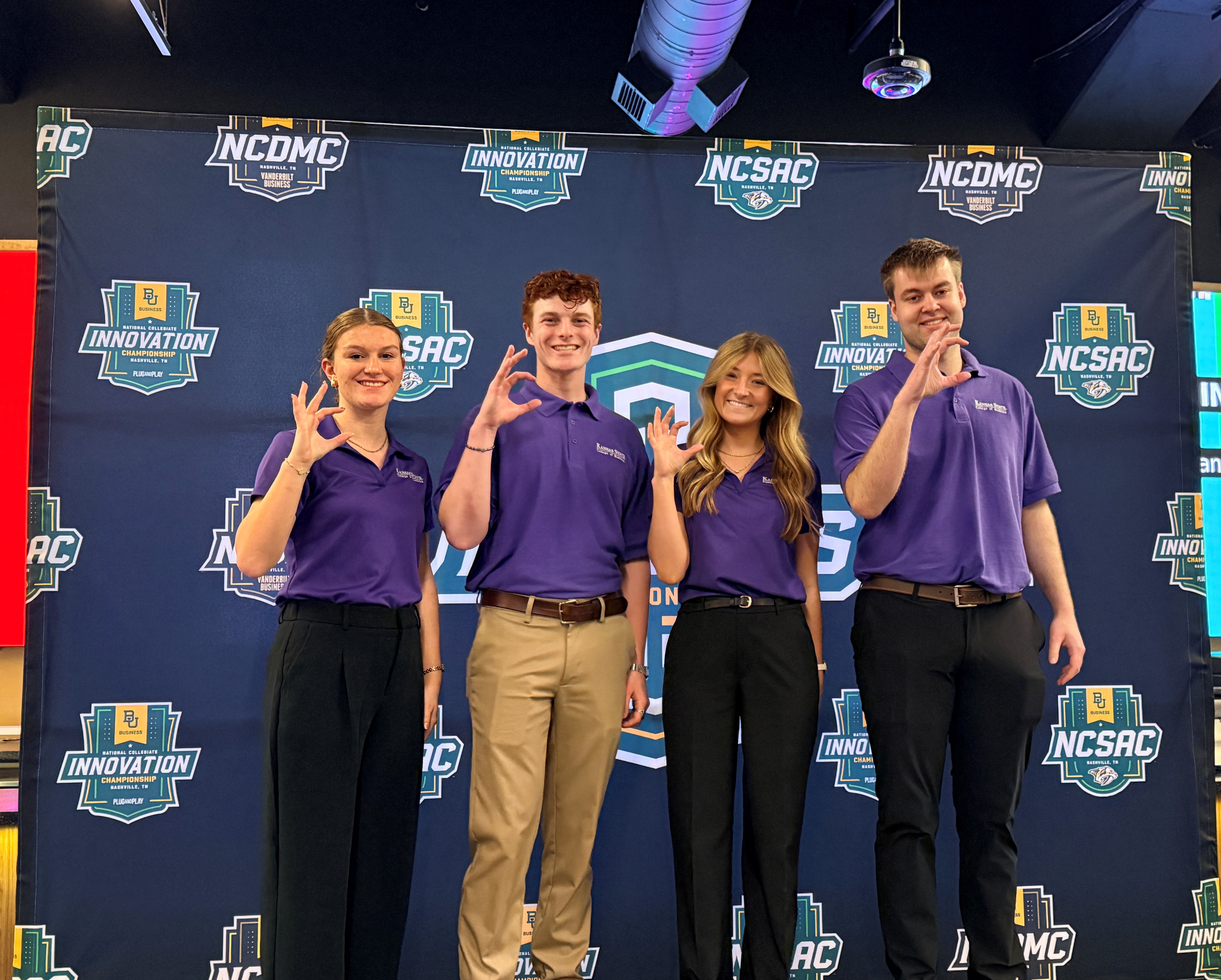 Four students stand in a line in front of a banner background for a marketing sales competition. The students are wearing purple K-State polos and giving the Wildcat hand sign.