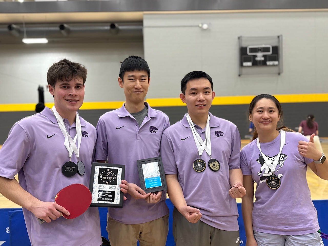 Four students wearing lavender K-State polos stand in a gym wearing medals and holding trophies. The students are on the table tennis team.