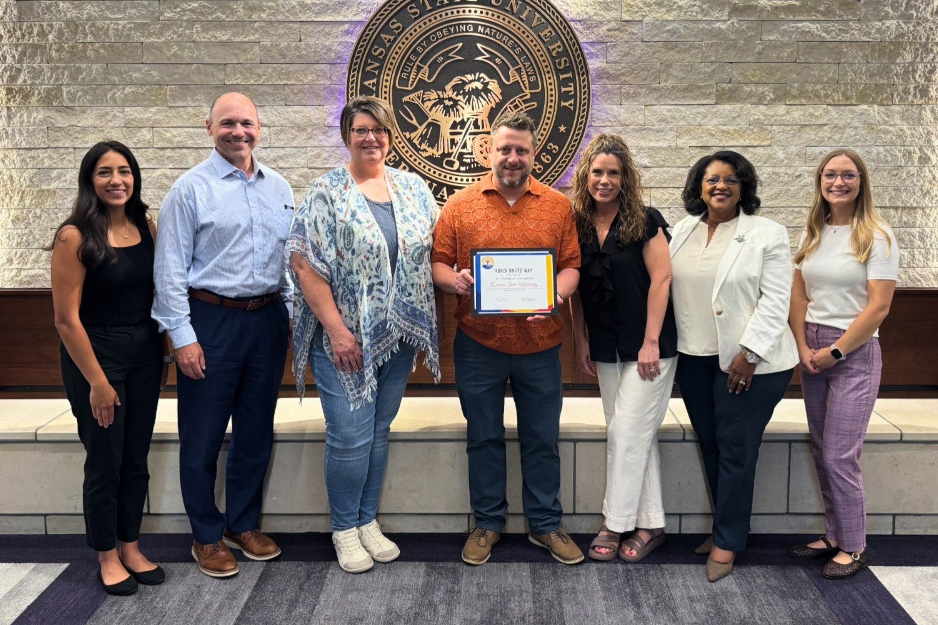Seven staff members stand in front of a purple K-State seal in Anderson Hall, holding the commemorative award received from Konza United Way