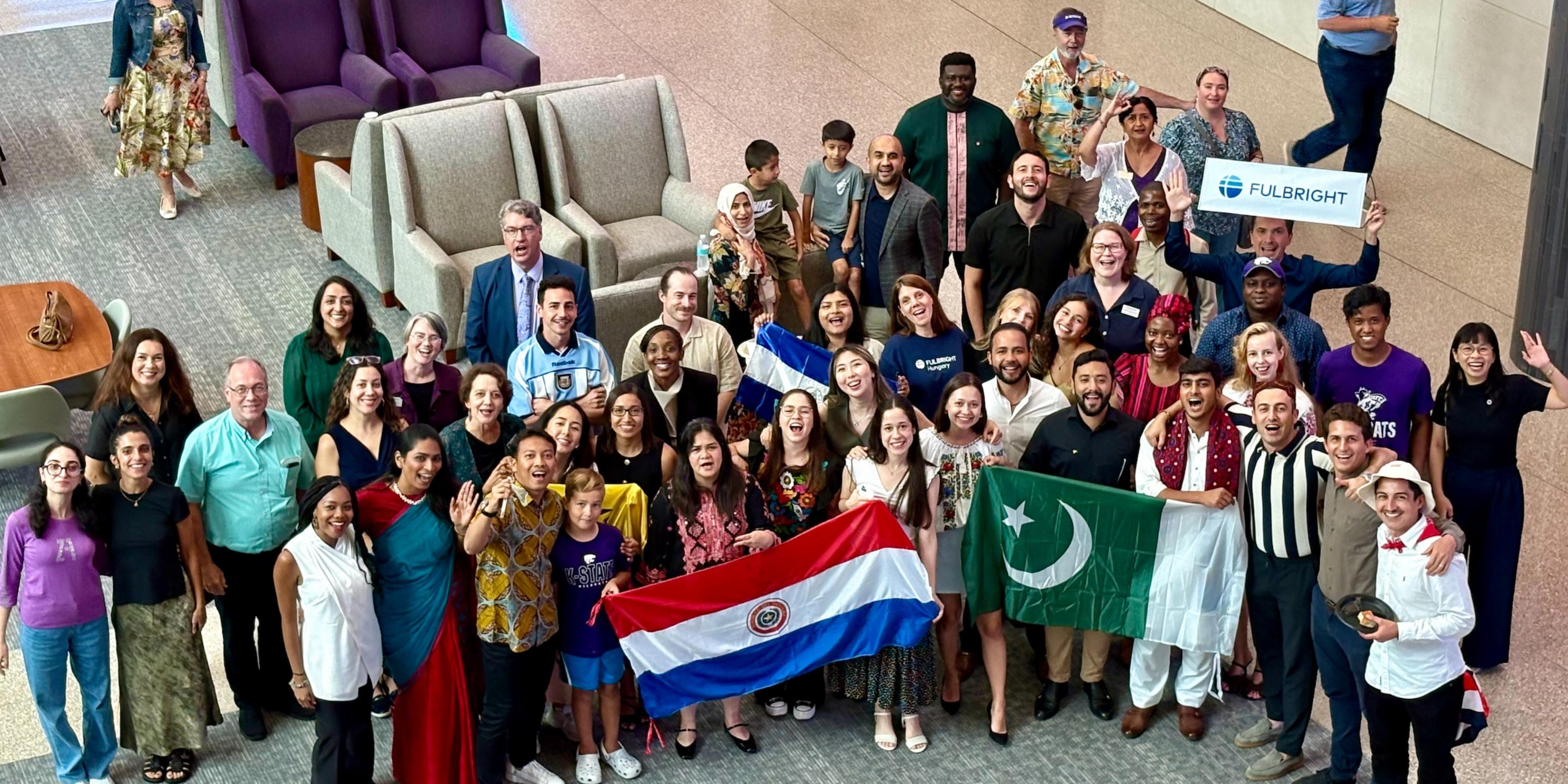 A large group of Fulbright students, holding flags from around the world, pose and smile for a photo in the College of Business atrium