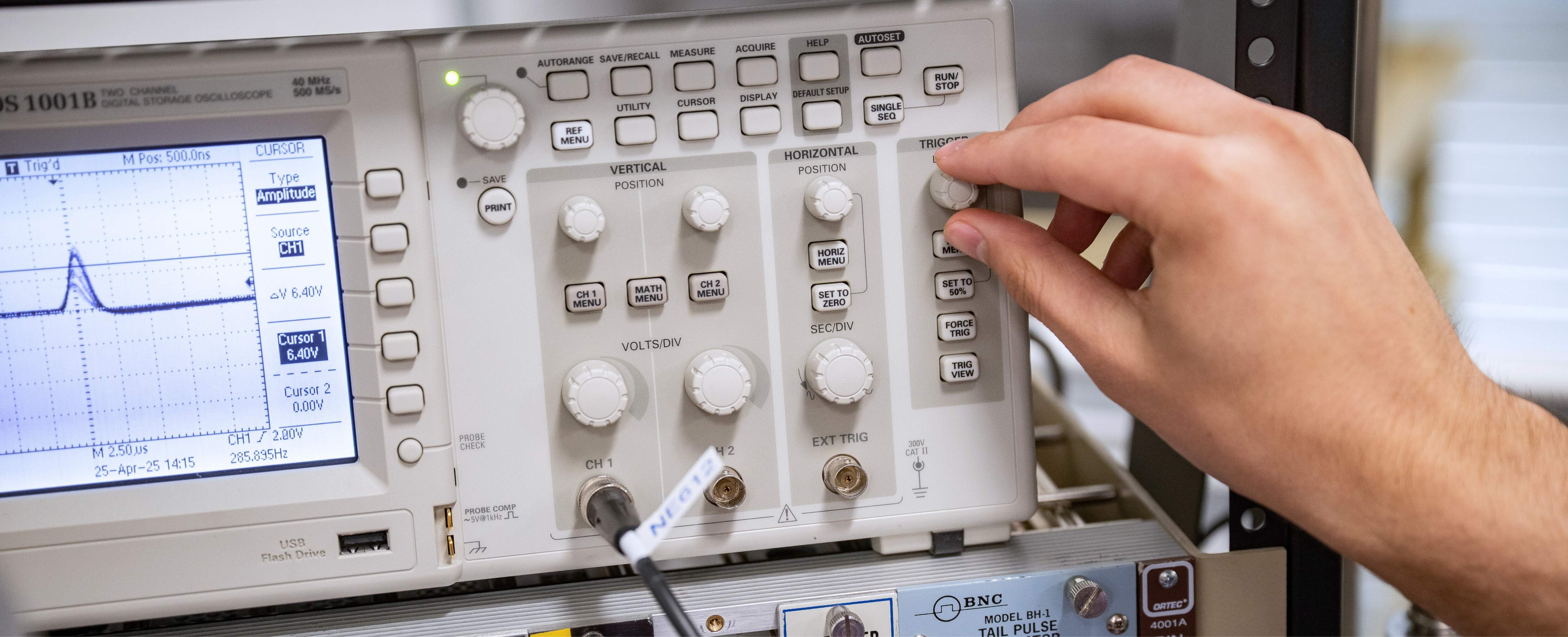 A hand reaches toward a knob on a research panel showcasing a variety of knobs, buttons and lights.