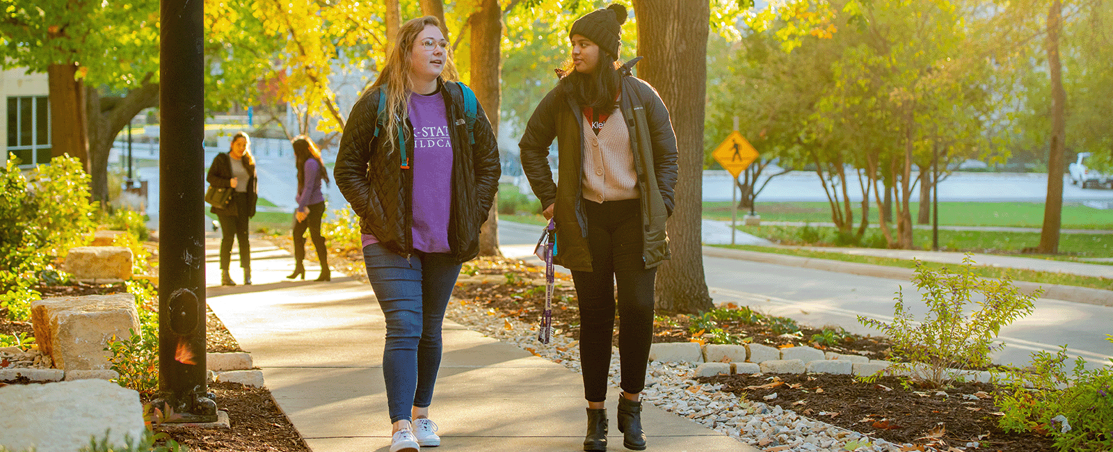 Students walk in front of Justin Hall. The sun lights up the foliage on the green trees.