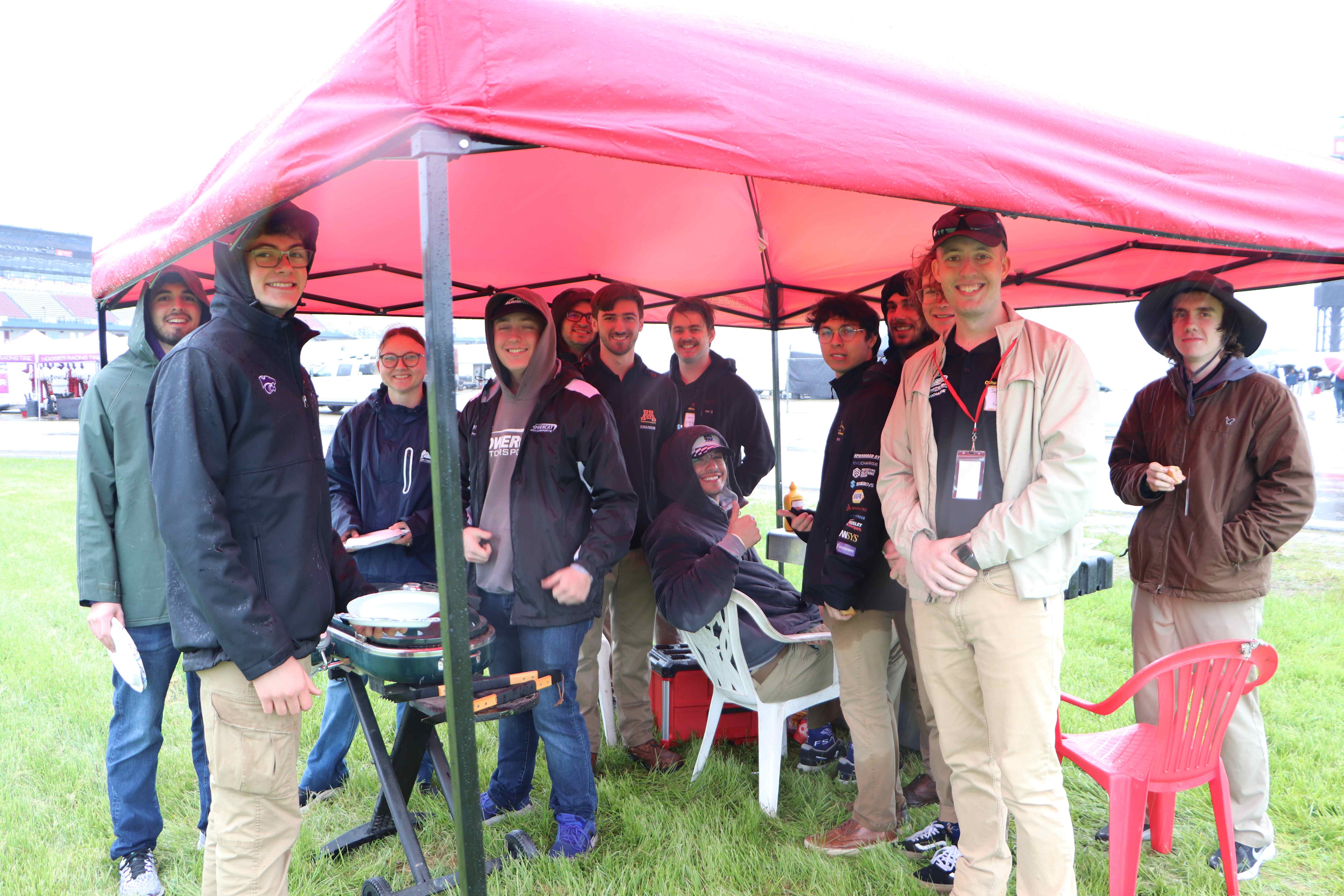 Some of our team under cover by our grill waiting for hot dogs made by our boy Ethan, some people from another team are present as well.