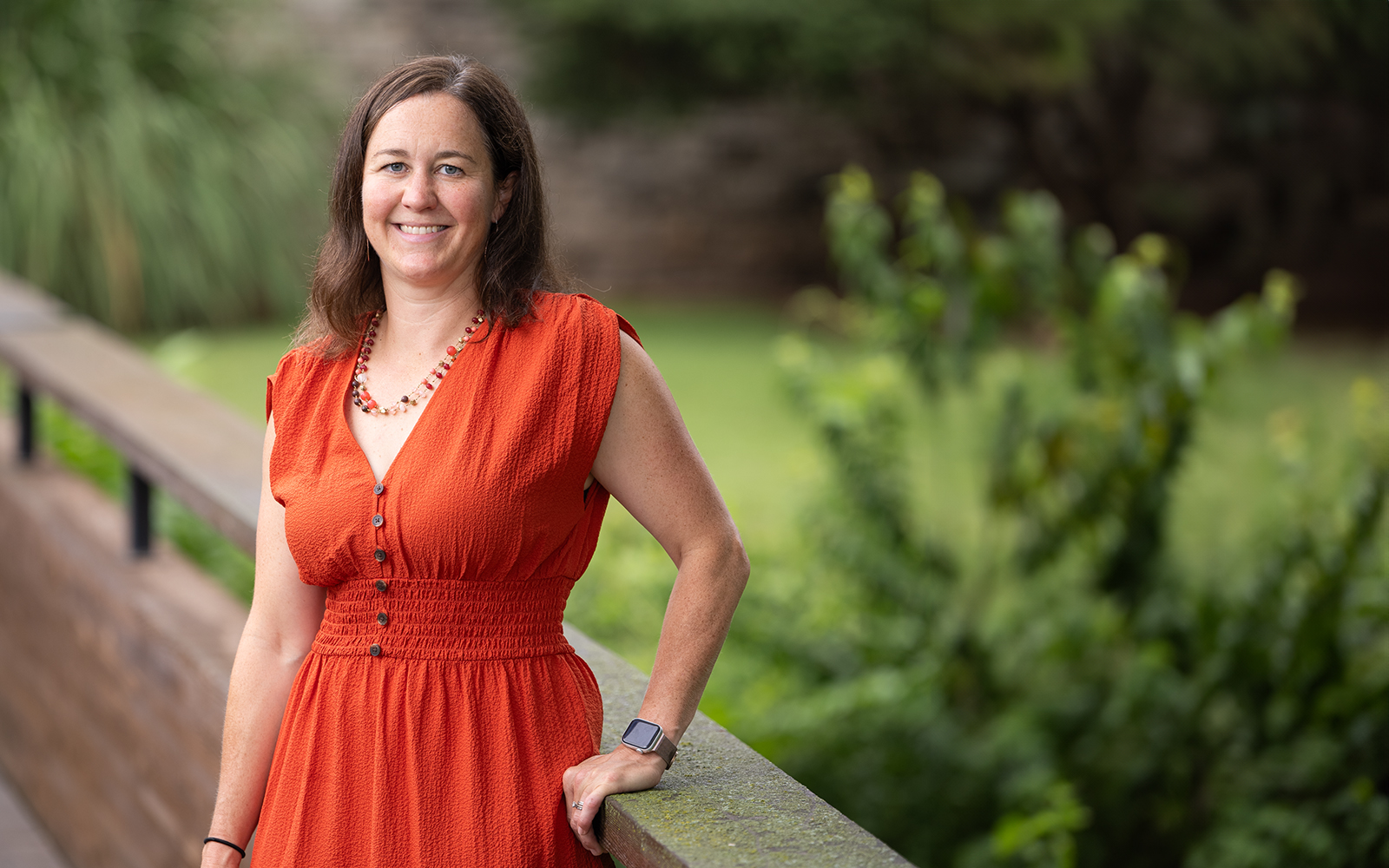 A woman in a red dress poses for a portrait on a bridge, with lots of greenery out of focus behind her.  