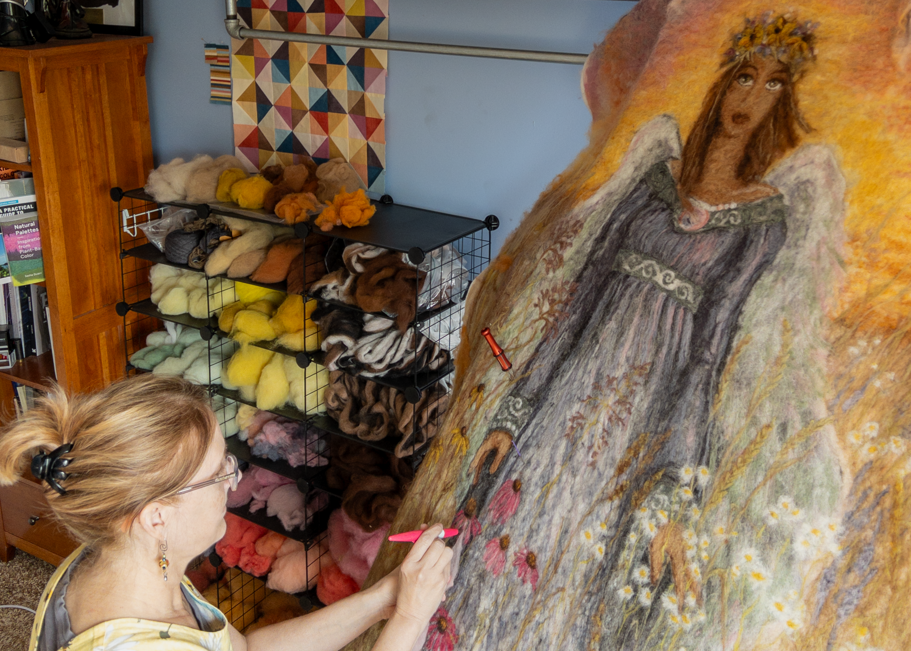 A woman pictured in front of a rack holding various colored fiber works on a felt coffin cover depicting a prairie angel.