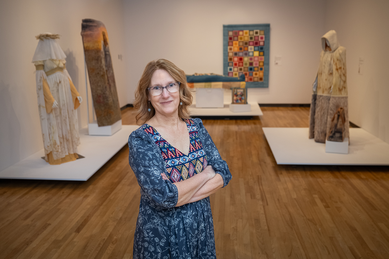 A woman in a blue patterned dress stands in front of an art museum exhibition featuring sustainable burial cloaks.