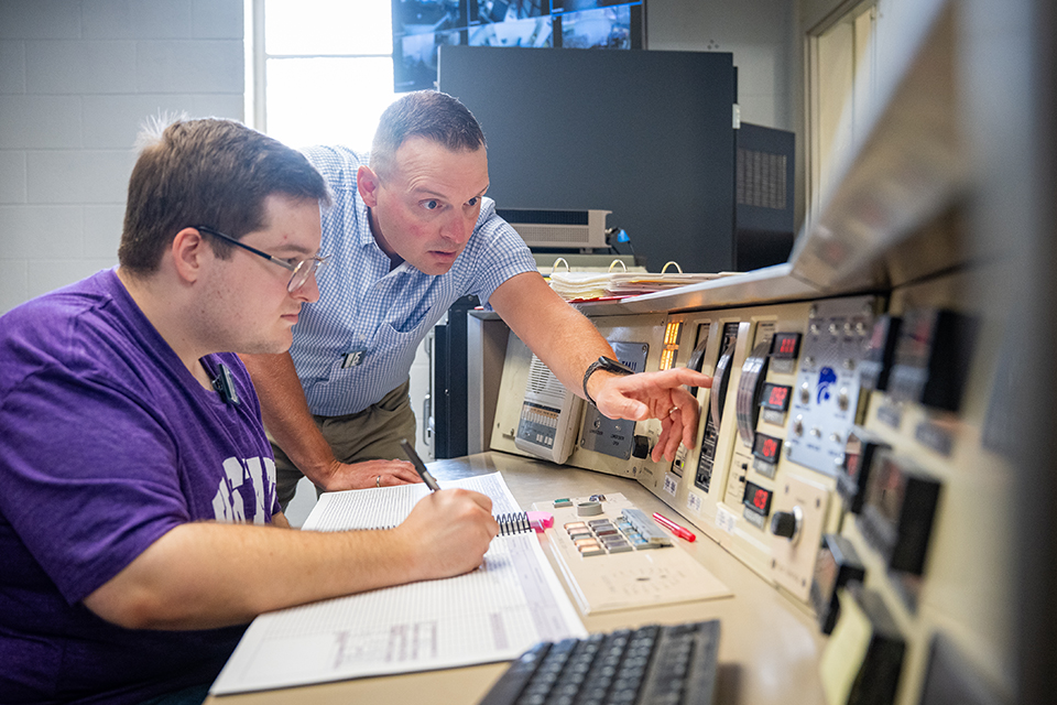 A student in a purple T-shirt sitting at a nuclear reactor control panel watches while a college professor points out one of the screens at the panel.