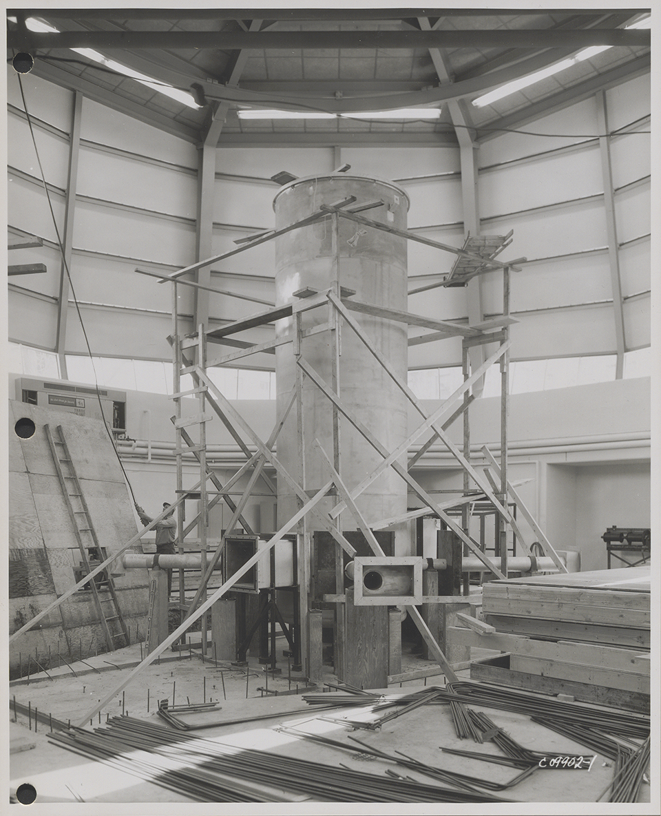 A black-and-white historical photo shows a tall nuclear reactor well under construction inside an even larger, cavernous indoor building.