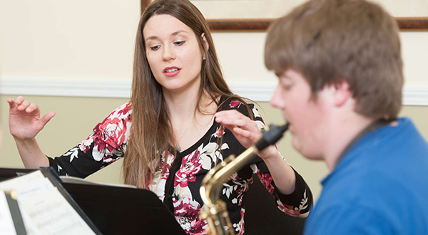Woman in patterned shirt conducts student in blue shirt playing saxophone.