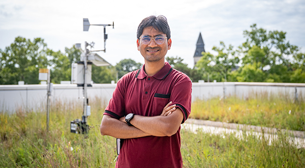A graduate student in a red polo stands with his arms crossed on a roof covered in tallgrass.
