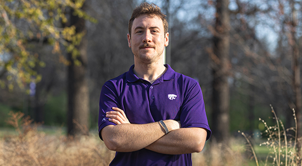 Student in purple shirt crosses arms and smiles for a portrait in wooded area.