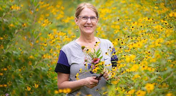A college professor holds a bunch of yellow flowers and poses for a portrait while standing in a bigger field of flowers.