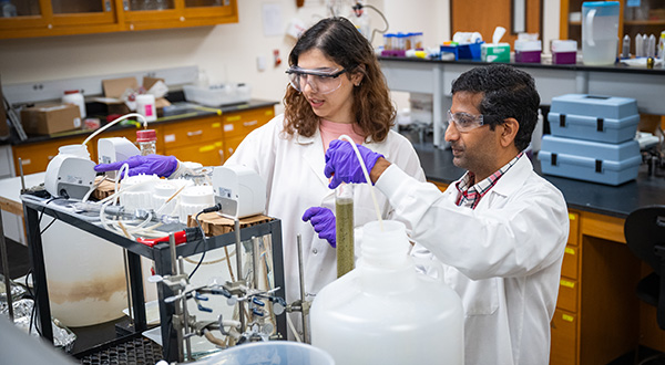 A man and woman in white lab coats with purple gloves stand in a lab and work on a project.