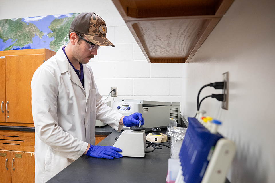 An undergraduate student researcher in a white labcoat works with equipment on a lab bench.
