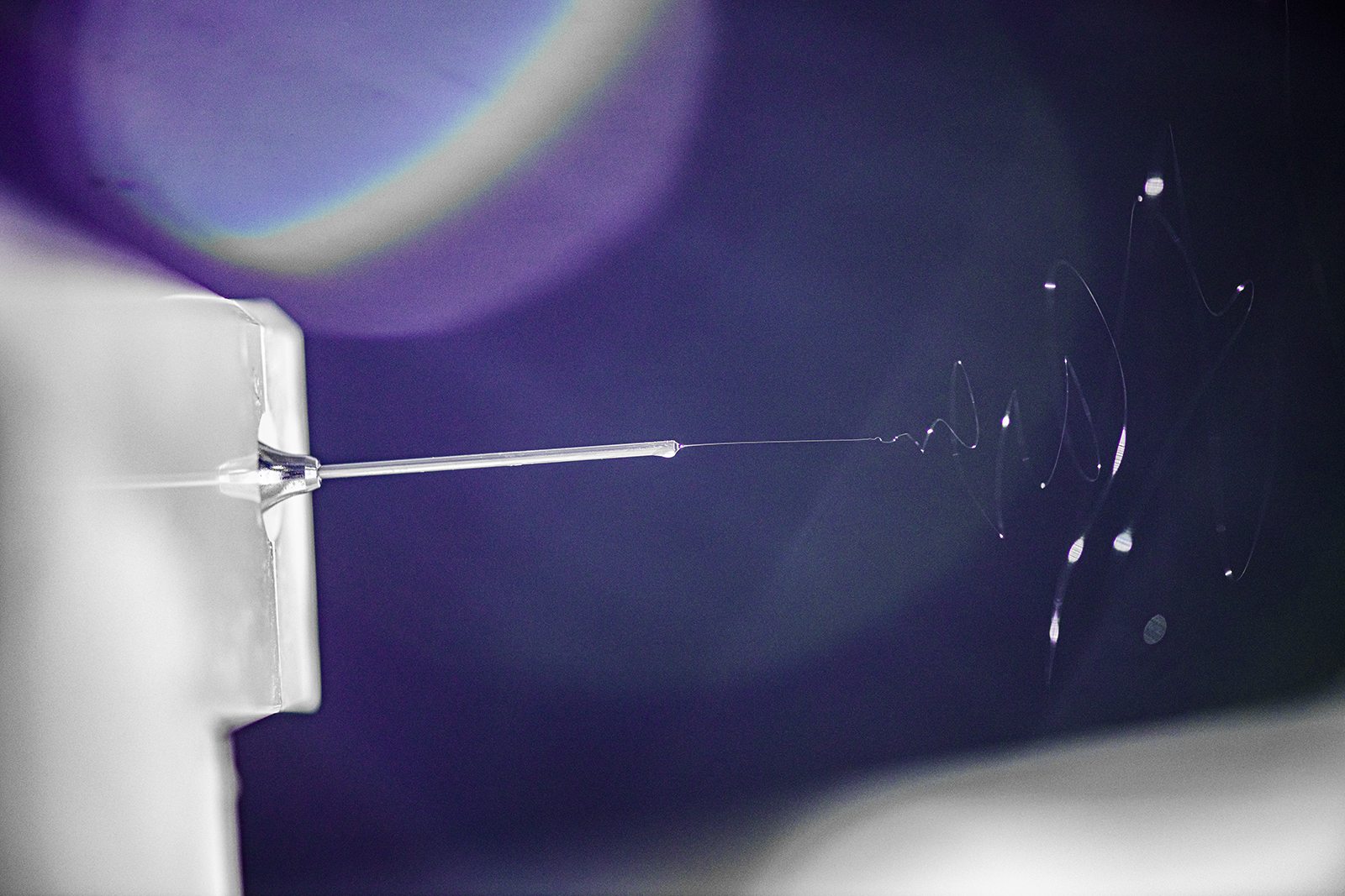 A microscopic image shows a swirl of fiber spinning out of a silver needle against a dark purple backround.